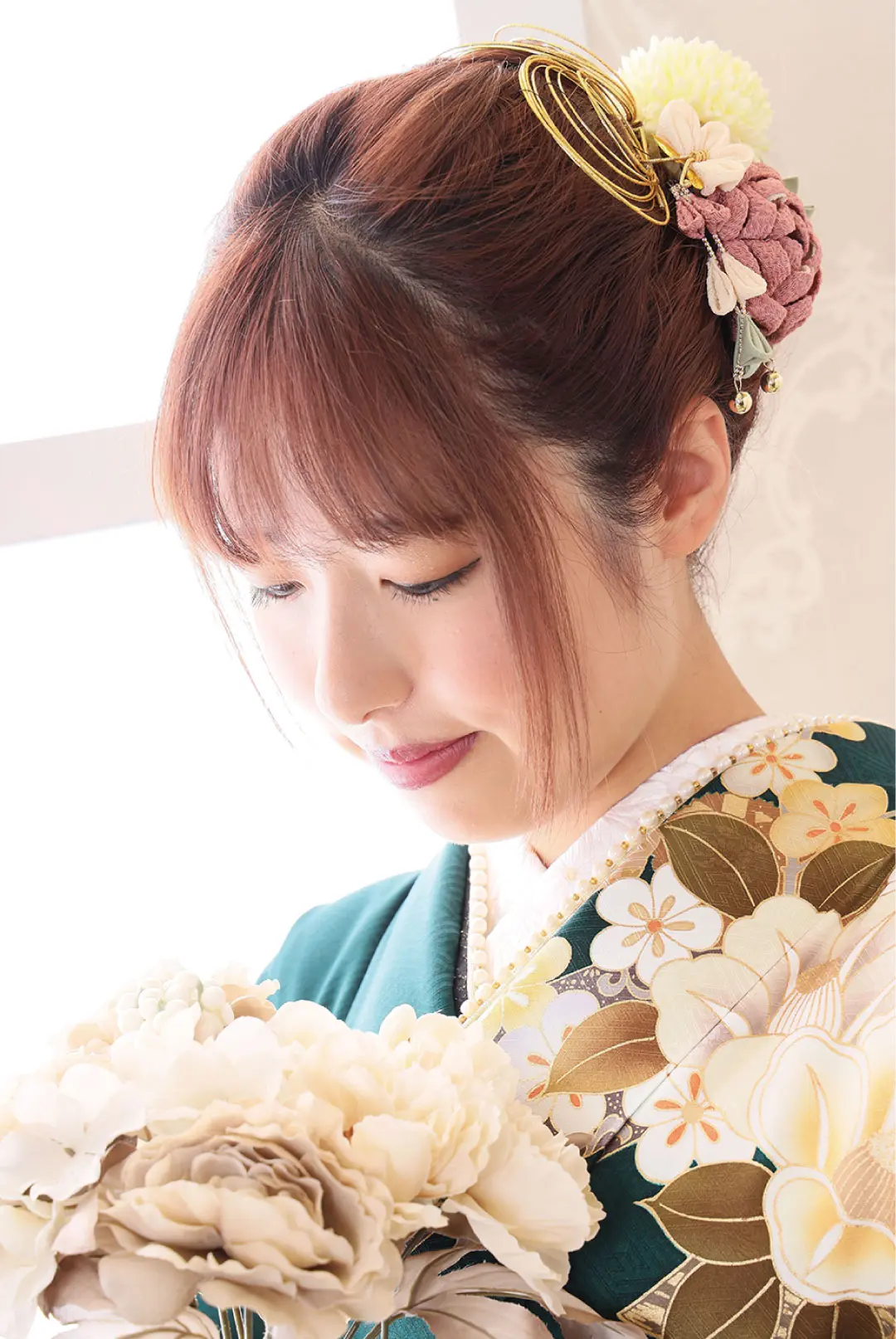 茶色の髪に花飾りをつけた女性が、金と白の花柄があしらわれた緑色の着物（令和6年成人式）を優雅に着こなしている。淡い花束を持ち、穏やかな表情でうつむいている。.
