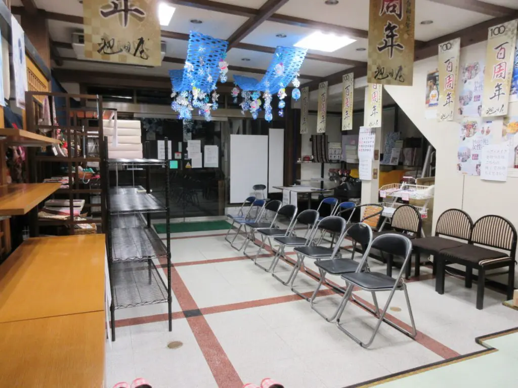 折りたたみ椅子が一列に並べられ、テーブルと棚が向かい合うように置かれた部屋の内部。天井からは紙で装飾された青い網が垂れ下がっている。壁には様々なポスターや看板が貼られ、床はタイルと堅木張りの模様になっている。.