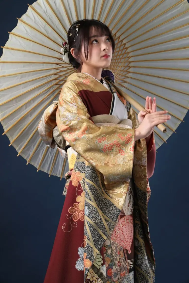 日本の伝統的な着物を着た人が、装飾的な日傘を肩にかけています。着物には金色とワインレッド色の複雑な模様が施されています。背景は濃い無地です。.