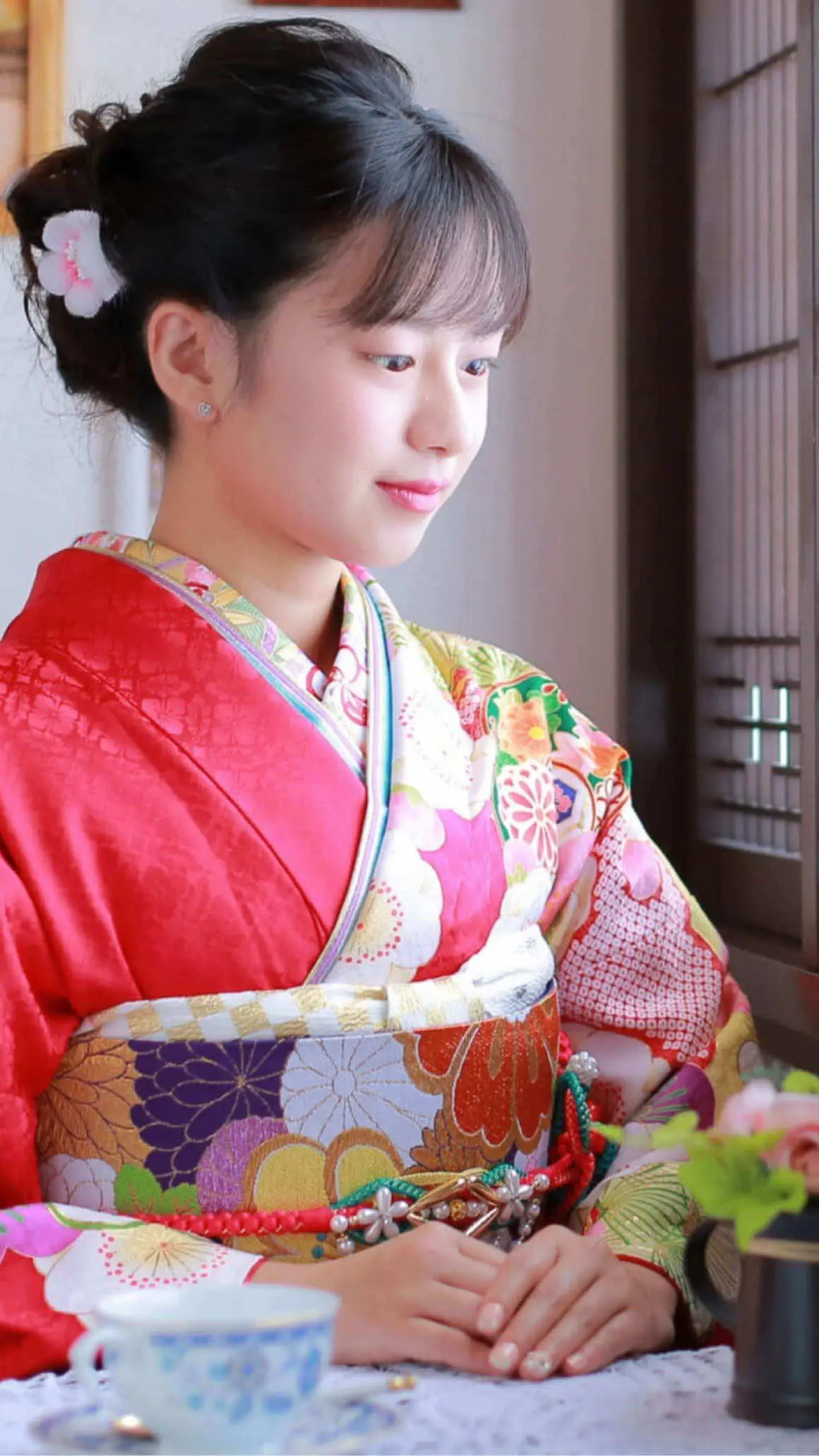 赤とピンクの鮮やかな花柄の着物を着た女性が窓辺に座っている。髪には白い花飾りが付けられている。彼女の目の前のテーブルには、ティーカップと花の入った小さな花瓶が置かれている。.