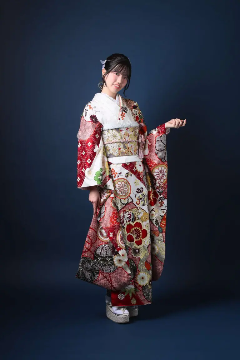 紺色の背景にポーズをとる女性。花柄や幾何学模様があしらわれたカラフルな着物姿を披露している。笑顔で腕を上げ、着物を引き立てている。髪にはエレガントなアクセサリーを飾っている。.