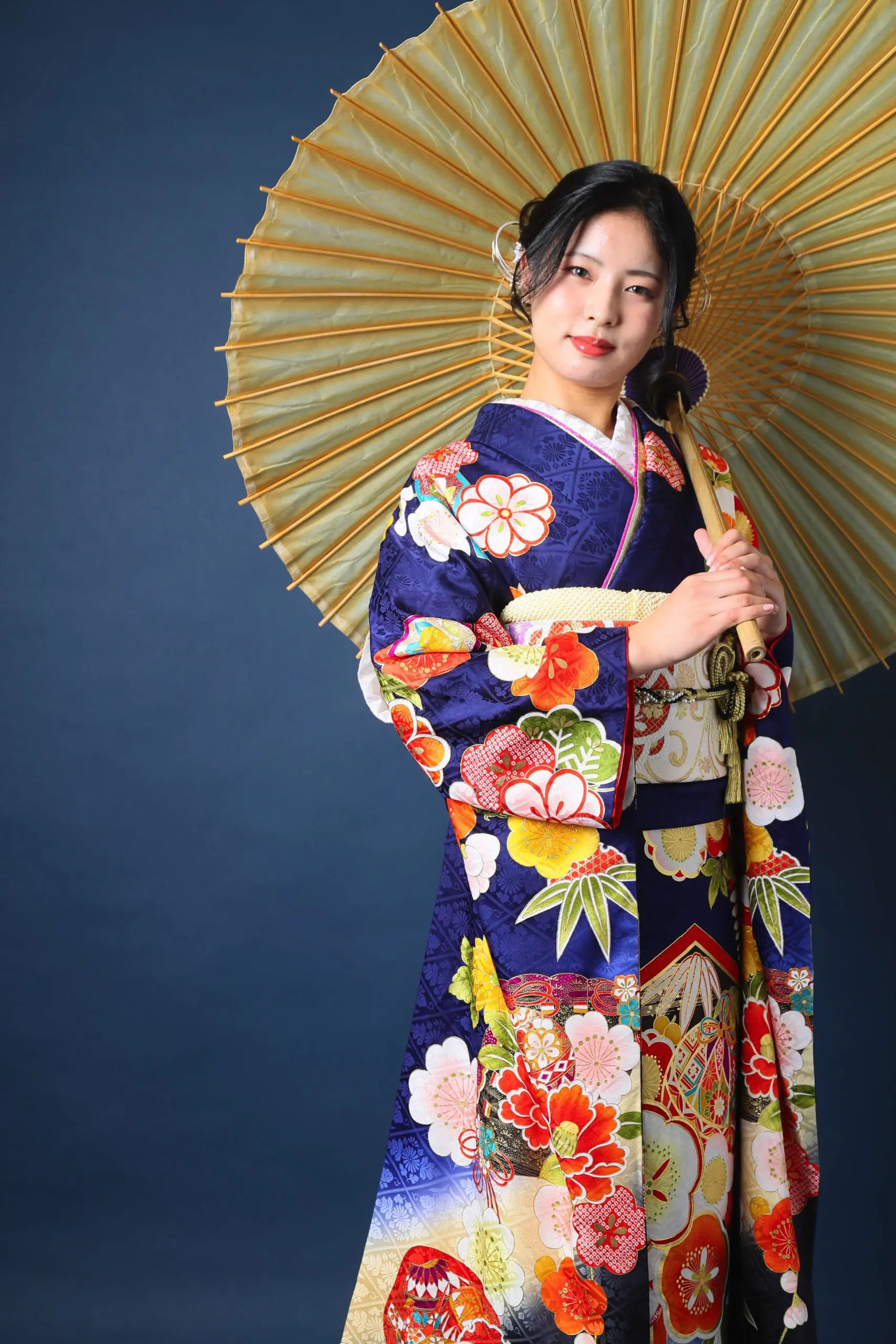 ダークブルーの背景に、鮮やかな花柄の着物を着た女性が伝統的な和紙傘を優雅に持っており、さいたま市の令和7年成人式のお祝いの精神を象徴しています。.