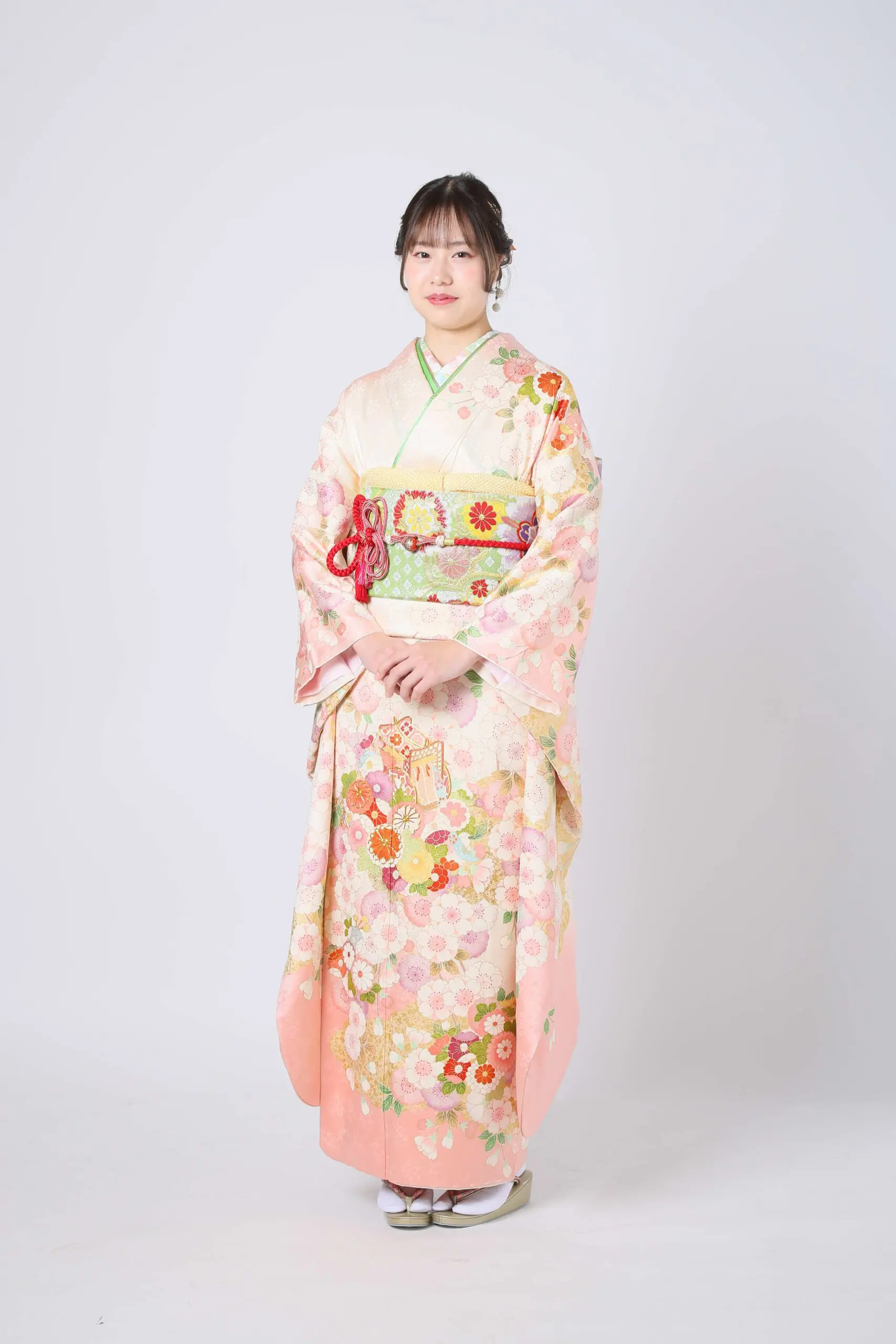 ピンクと花柄の着物に、幅広で色鮮やかな帯を締めた人物が、無地の背景に立っています。黒髪はゆるくまとめられ、両手は優しく前で組まれています。.