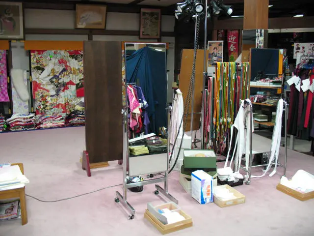 全身鏡のある部屋。周囲には色鮮やかな着物や帯などの伝統的な衣装が飾られ、床には箱や衣類が散らばっている。試着室や着付け室のような空間だ。.