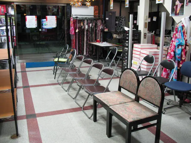 店内には椅子がずらりと並べられている。金属製の折りたたみ椅子、クッション付きの椅子、テーブルが置かれている。背景には、色とりどりの衣服が置かれたラックと、箱が置かれた棚が見える。床はグレーと赤のタイル張りだ。.