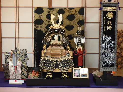 金と黒の細工が施された武士の甲冑が、装飾屏風を背に誇らしげに飾られています。その周囲には、包装された贈り物、幟、そして二人の肖像画が描かれた小さな額など、儀式に使われる品々が飾られています。精巧な意匠は、まるで毎日、空気中に響き渡る物語を物語っているかのようです。.