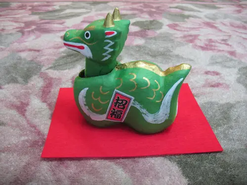 白と金の装飾で飾られた小さな緑色のセラミックの龍の置物が赤い布の上に置かれています。大きな目と二本の角が愛らしく、「ありがとうございました」の赤と白の文字が刻まれています。背景は花柄です。.