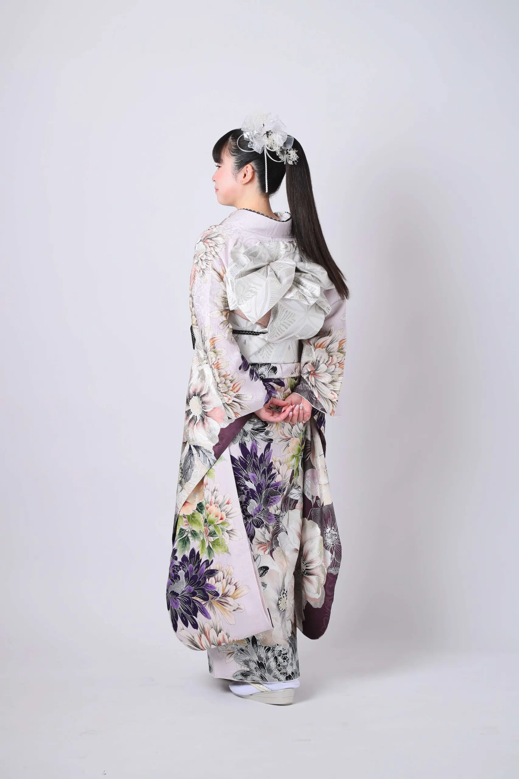 花柄の伝統的な着物と大きな銀色の帯を締めた人物が、両手を後ろで組んで立っています。カメラから背を向け、髪には白いアクセサリーを飾っています。.
