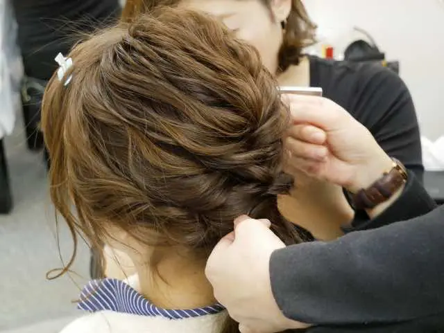 茶色の髪の人物が、別の人物に髪を三つ編みにしてもらっている。この場面は屋内で撮影されたもので、人物の後ろ姿と横顔しか映っていない。.