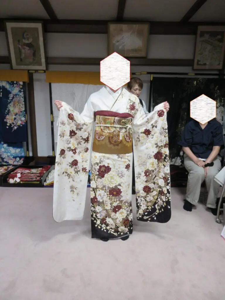 花柄と華やかな帯を締めた日本の伝統的な着物を着た人物が、室内で振袖展に参加している。近くには二人の人物が座っている。顔は隠されている。室内の背景には、額装された美術品や衣装の展示が見られる。.