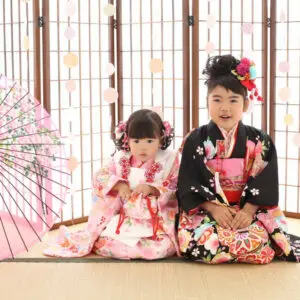 色鮮やかな日本の伝統的な着物を着た2人の幼い子供が、木製の屏風の前の畳の上に並んで座っており、左側には装飾的な傘が置いてあります。.