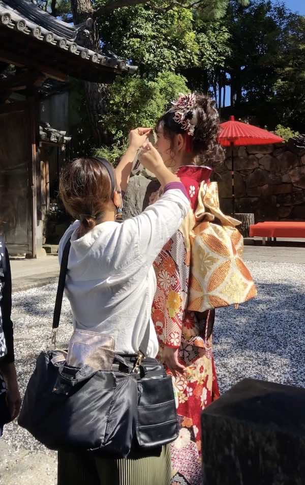 白いトップスを着た女性が、屋外の石垣と赤い紙傘の近くの場所で、装飾的な帯を締めた日本の伝統的な着物を着た別の女性の髪を整えている。.